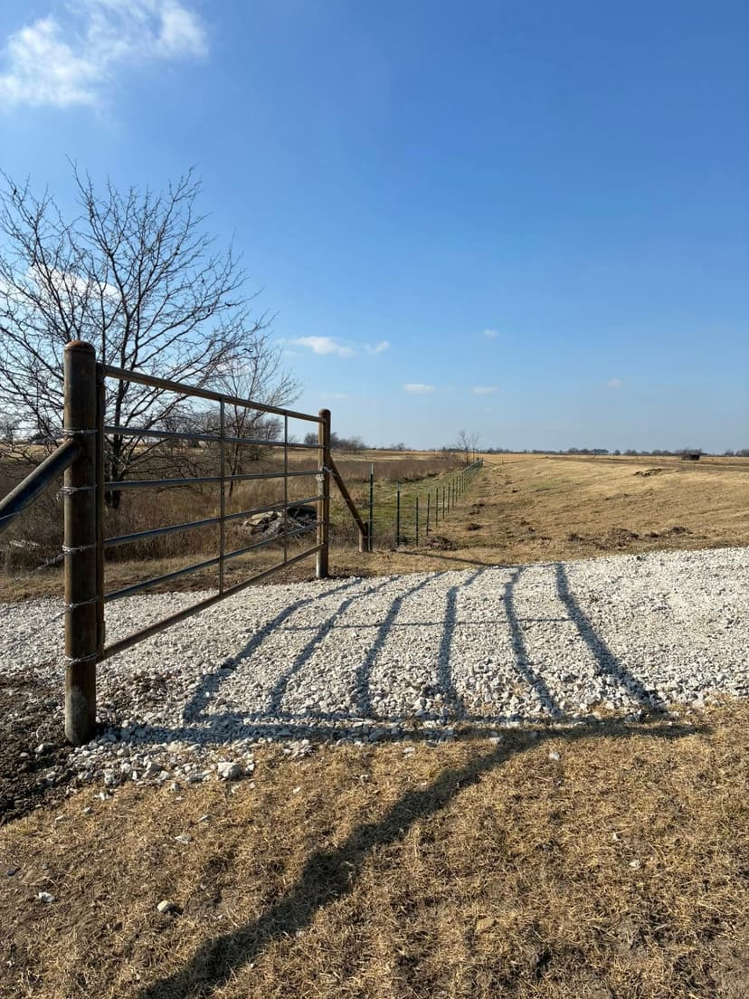 Rustic metal gate on gravel path, leading to open field under a clear blue sky.